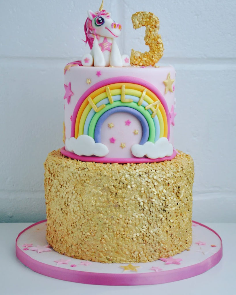 a two tiered gold, white and rainbow cake with a unicorn on top