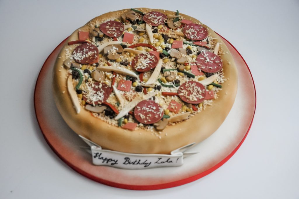 A cake in the shape of a pizza and decorated liked one too with realistic pepperoni, chicken strips, sweetcorn, mushrooms and peppers