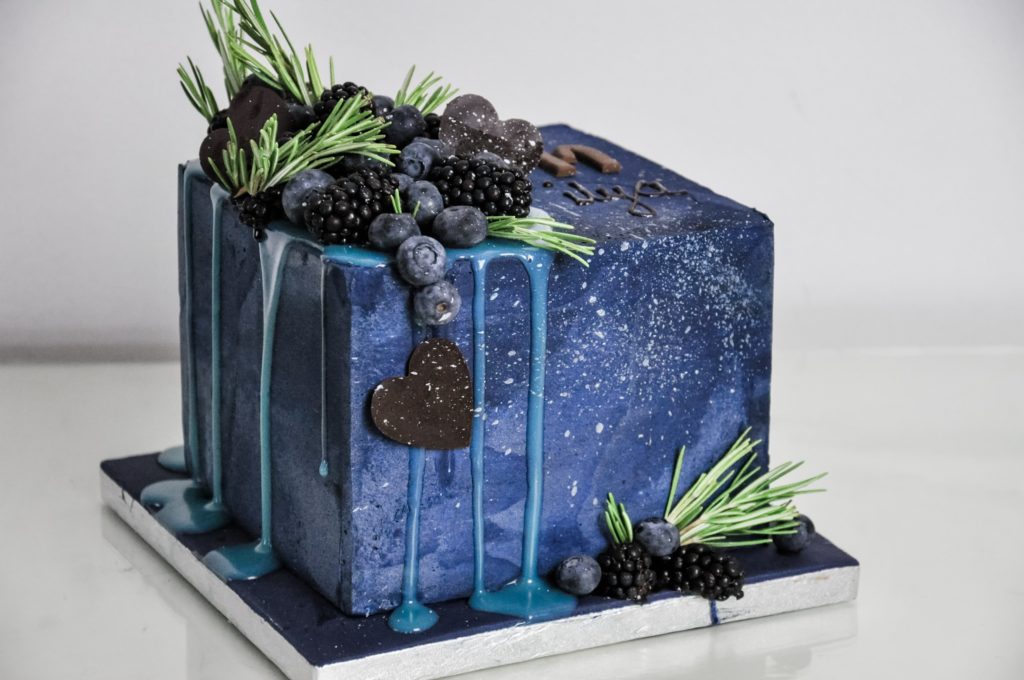 A square midnight blue buttercream cake with blue drip, dark chocolate hearts, rosemary, blueberries and blackberries on top