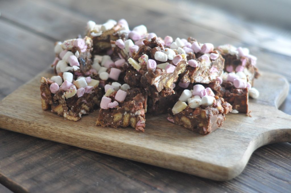 Squares of rocky road with pink and white marshmallows chopped up and piled high on a wooden chopping board